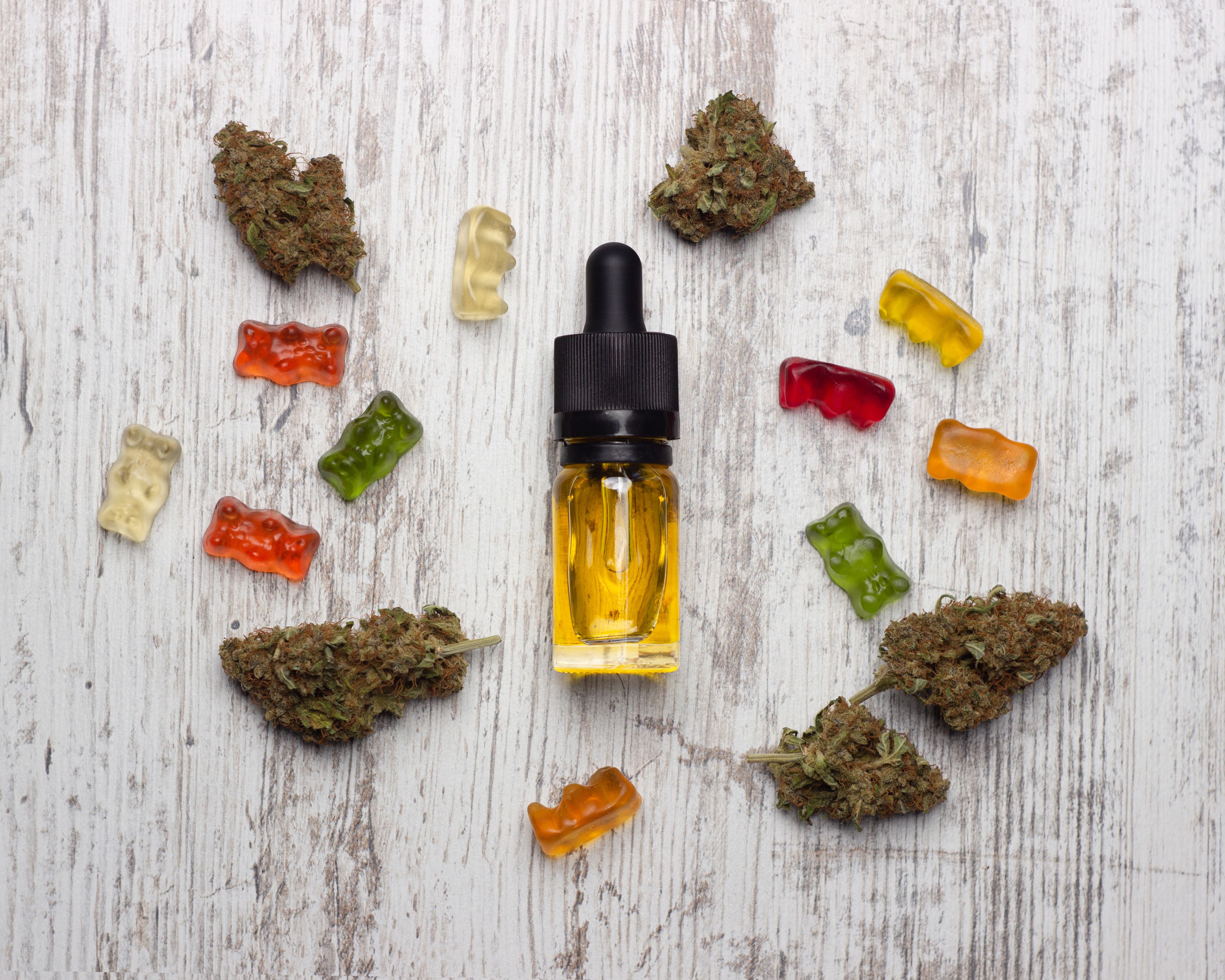 Gummies and cannabis buds lay spread out around a tincture CBD bottle on a white wood background.