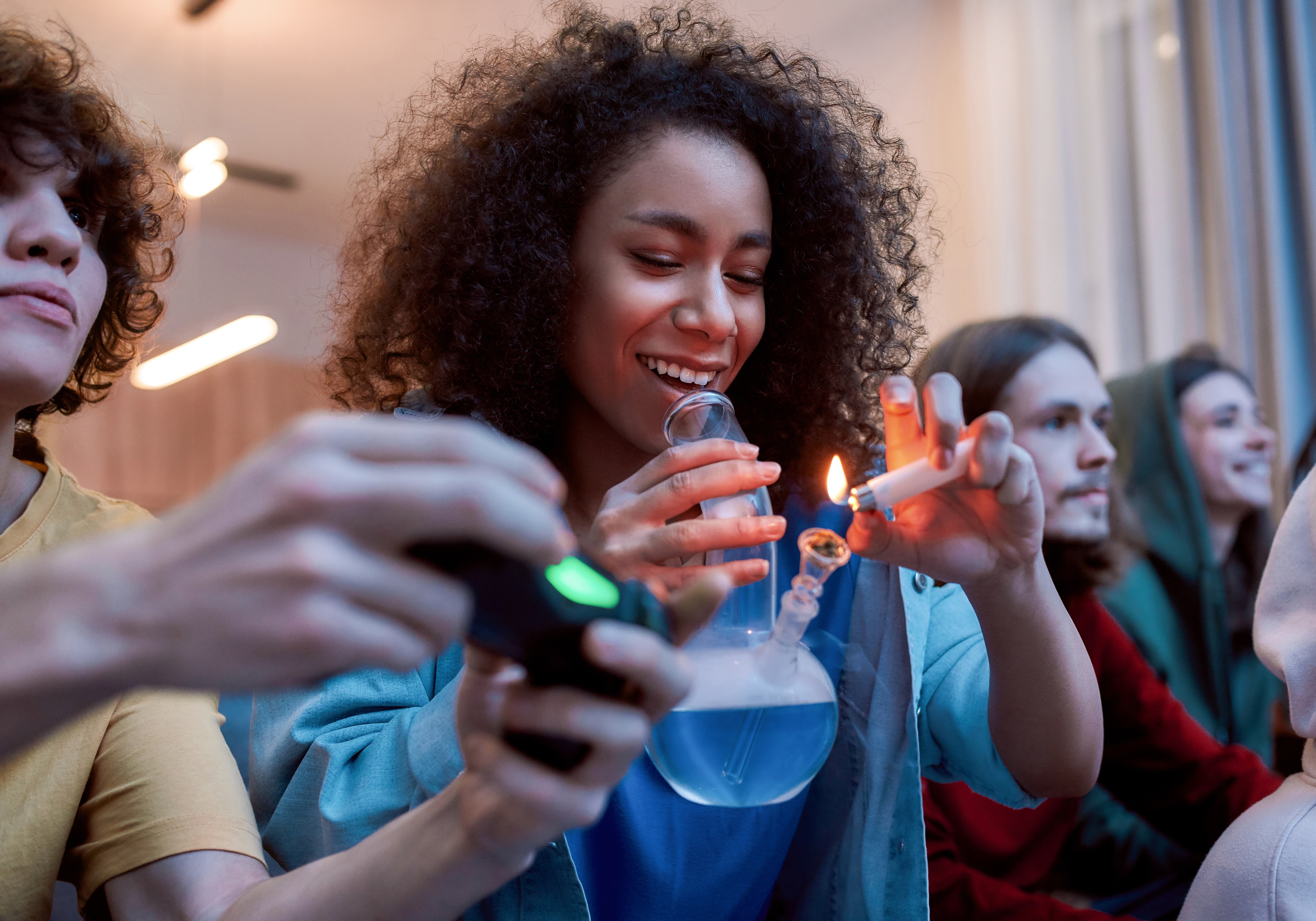 A young woman lights a bong while friends next to her play video games.