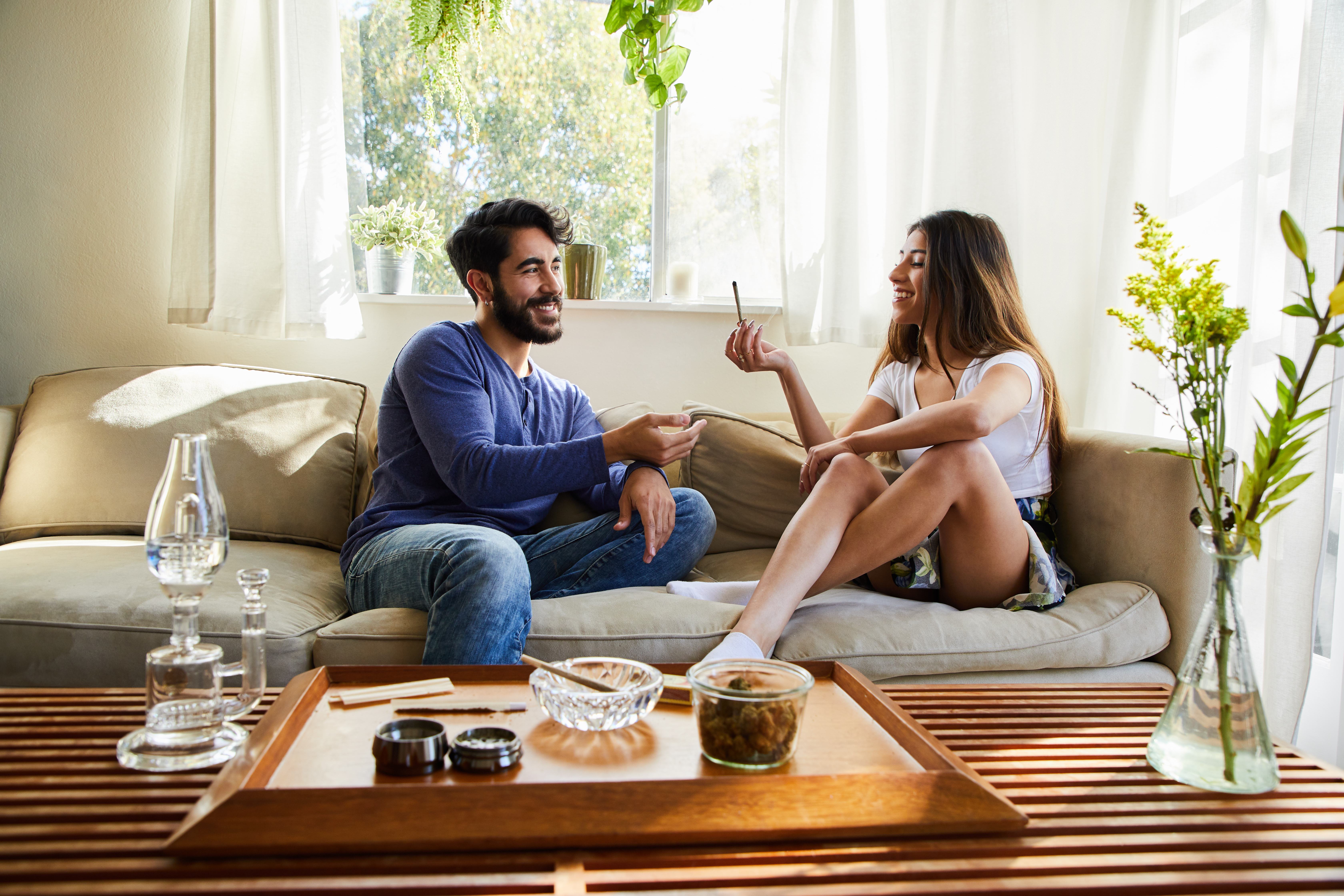 A young couple sit on a couch at their home smiling and smoking a joint.