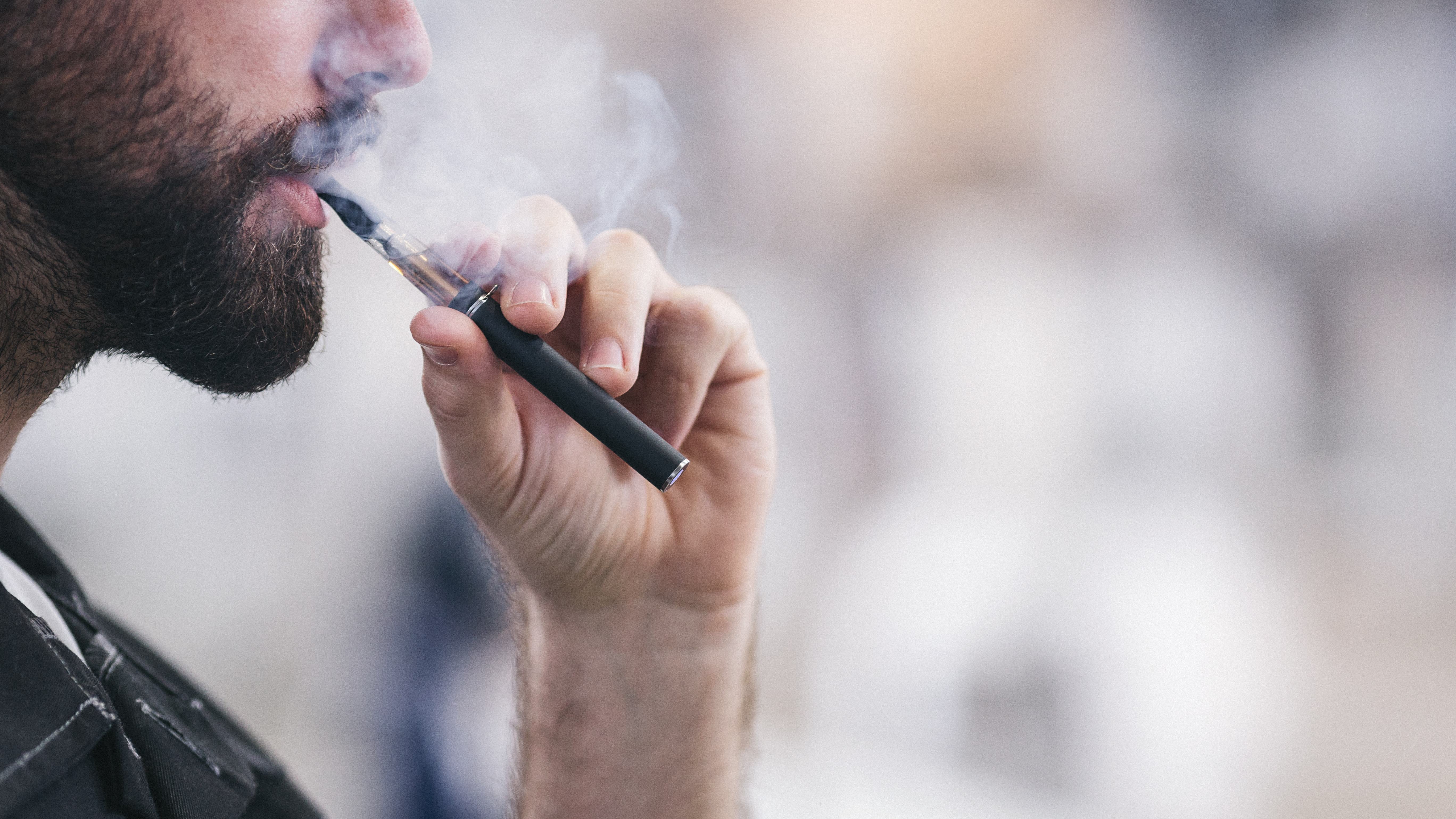 Bearded man taking a puff from vaporizer pen with a cloud of smoke.