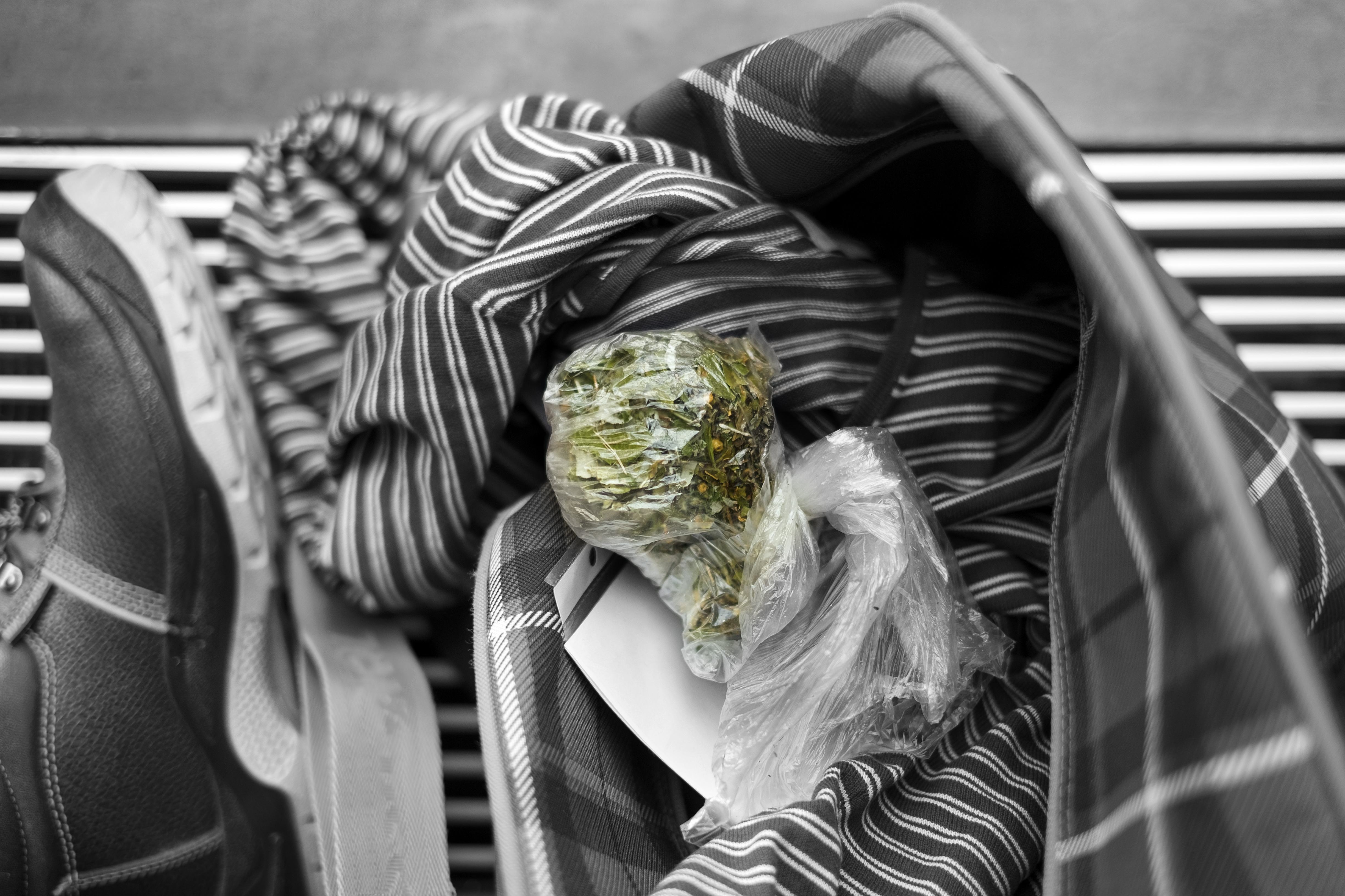 A bag of cannabis lays on top of black and gray colored clothes in an open suitcase.