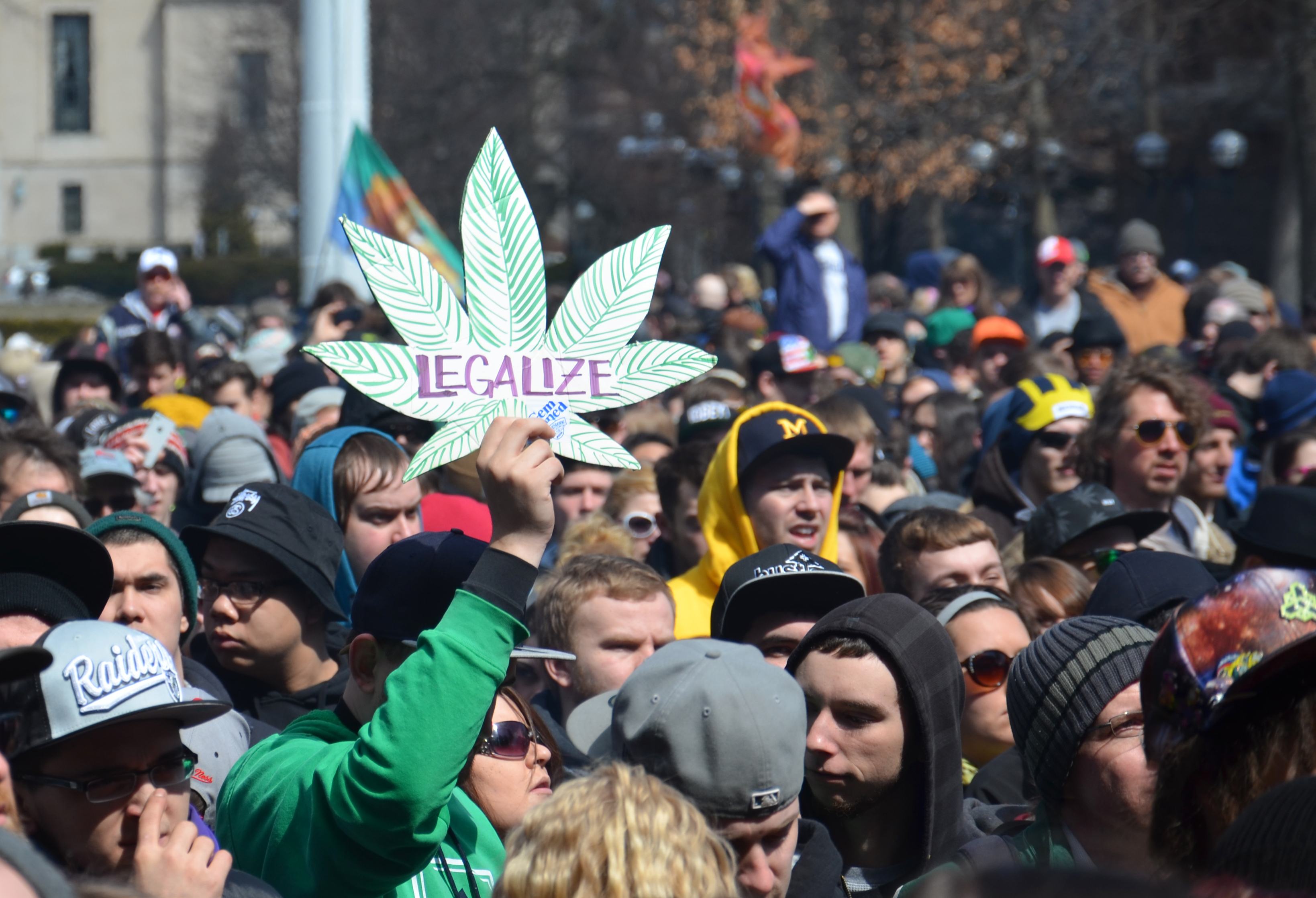 A large group of people are gathered with one person holding up a Marijuana leaf sign that says Legalize.