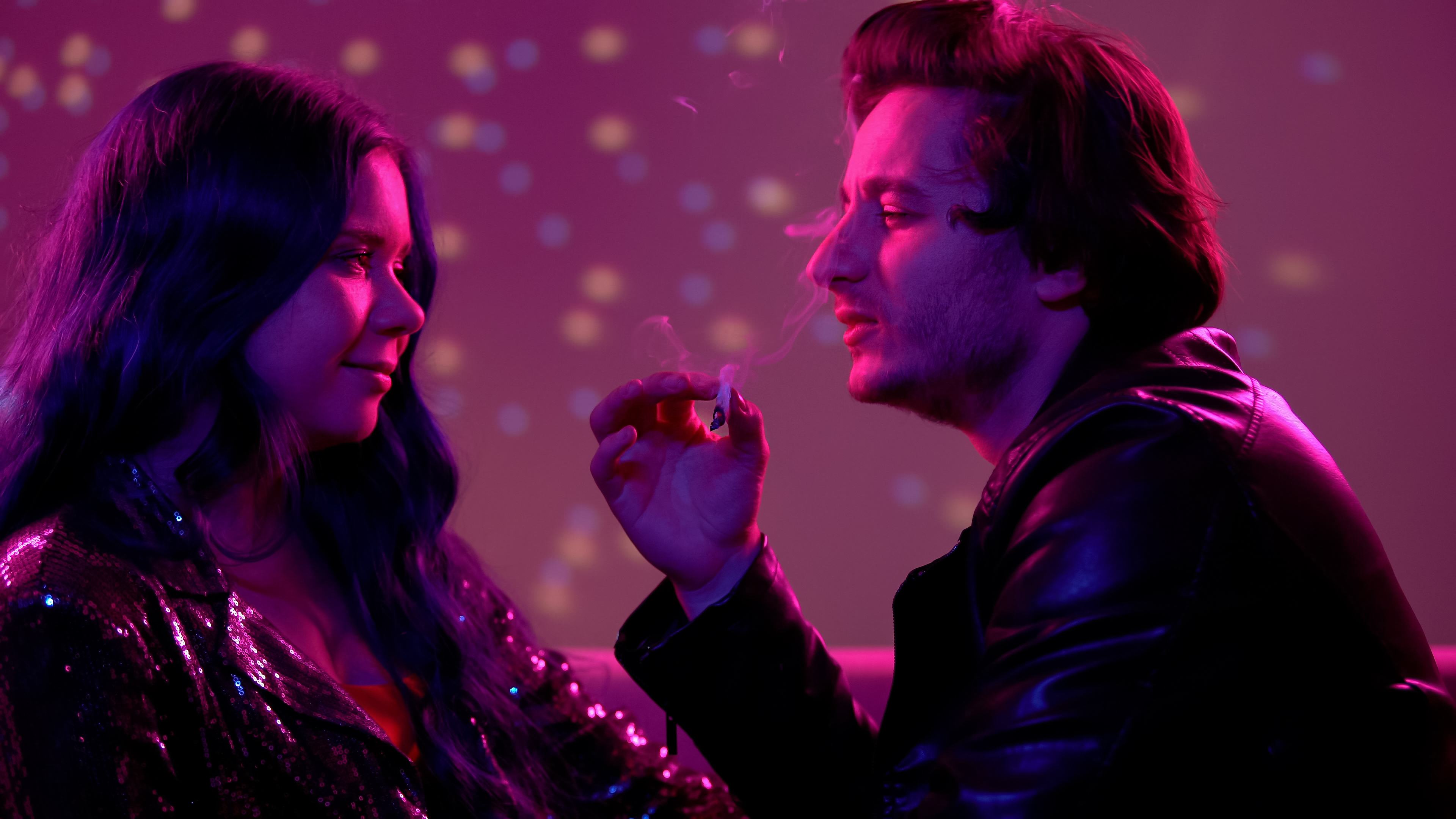 A couple face each other while at a club with the man holding a joint in his hand. The background is dark purple.