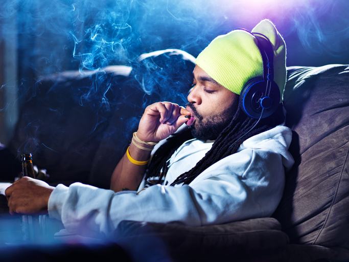 Man sitting on the couch, smoking a joint with headphones on and a beer in his hand.