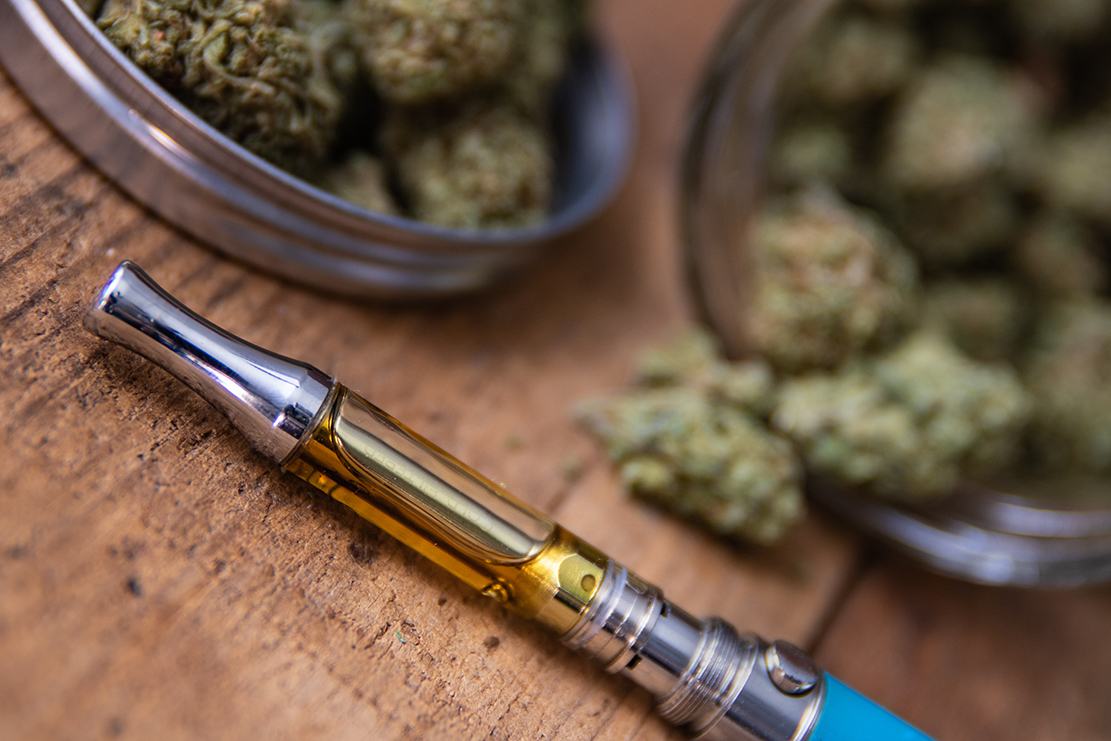 A Teal Blue Vape pen lays next to cannabis flower placed in silver tin cans on a wooden desk.